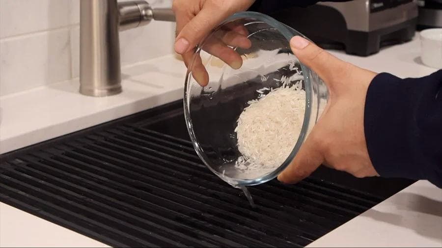 Rinsing uncooked white rice in a glass bowl over a black sink mat in a kitchen.