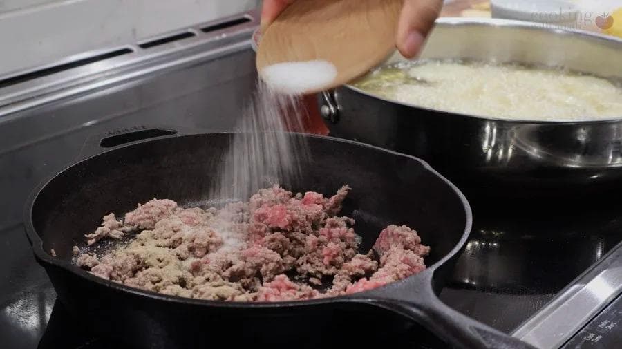 Salt being sprinkled into a skillet of browned ground meat and pork mixture during preparation of Perkedel Daging Kentang.