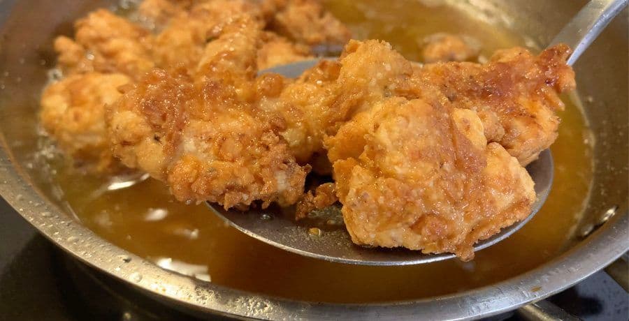 Golden-brown fried chicken pieces being lifted from hot oil with a slotted spoon in a stainless steel pot.