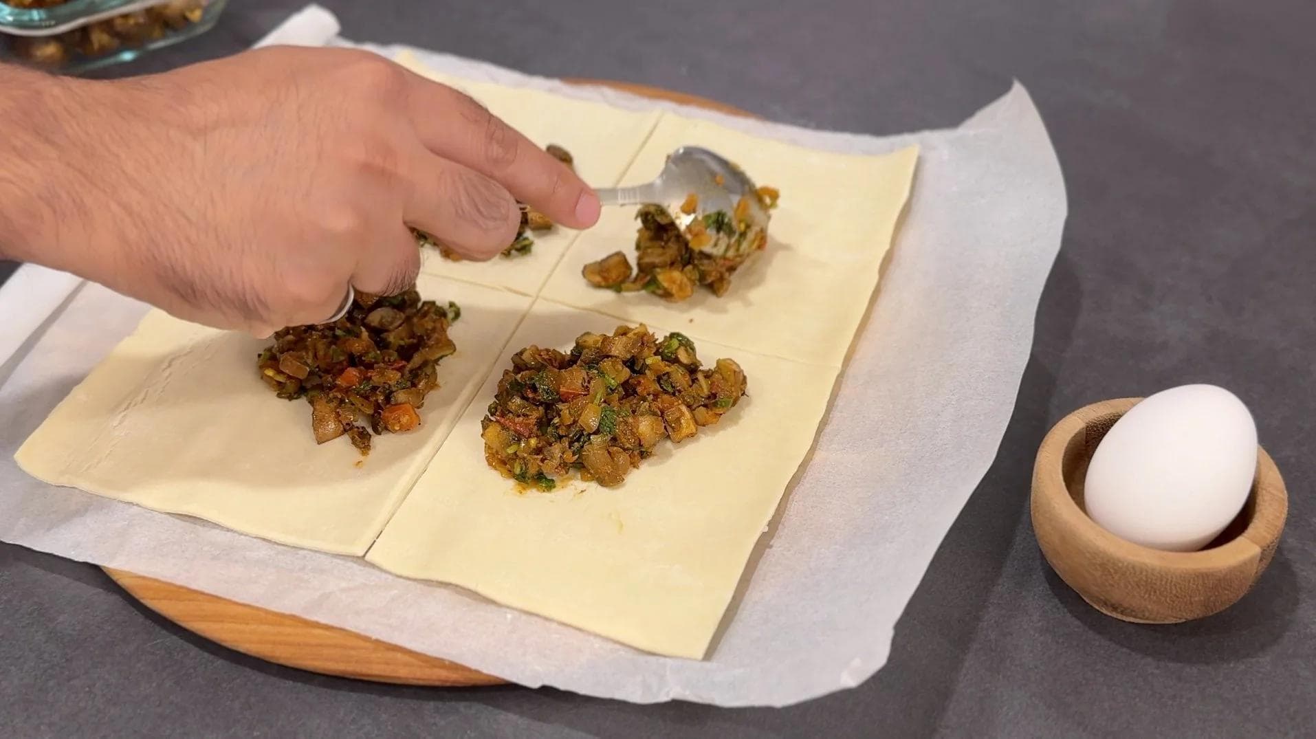 Hand filling puff pastry squares with mushroom mixture on parchment paper, with egg wash and wooden egg holder nearby.