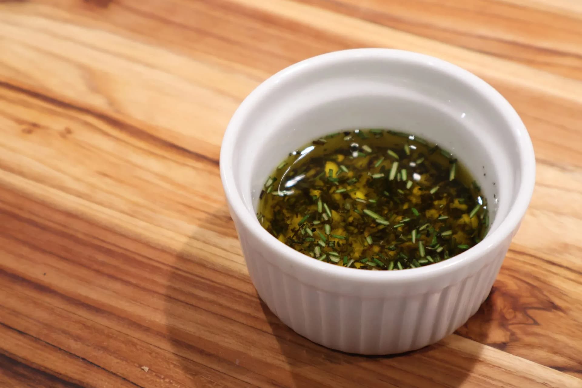 Small white ramekin filled with golden herb-infused garlic oil, sitting on a wooden cutting board.
