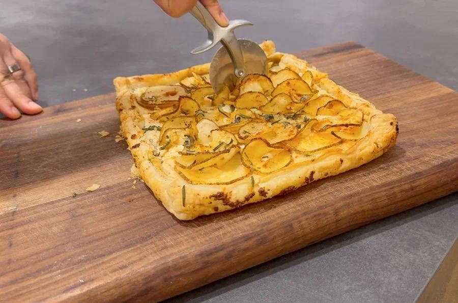 Pizza cutter slicing a golden baked puff pastry tart topped with thinly sliced potatoes and herbs.