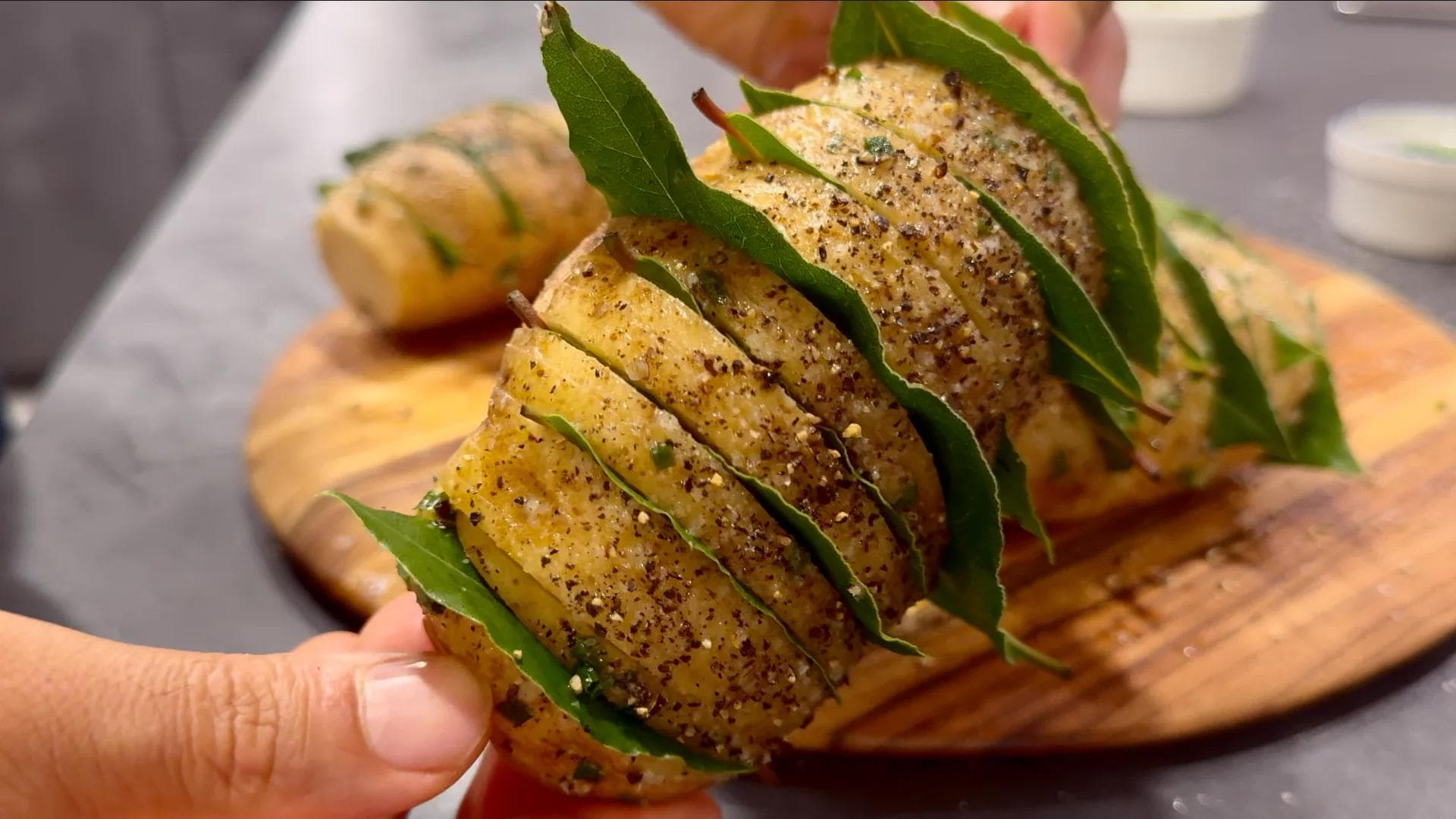 Hasselback potatoes with bay leaves inserted between slices, brushed with oil and seasoned with black pepper on a wooden board.