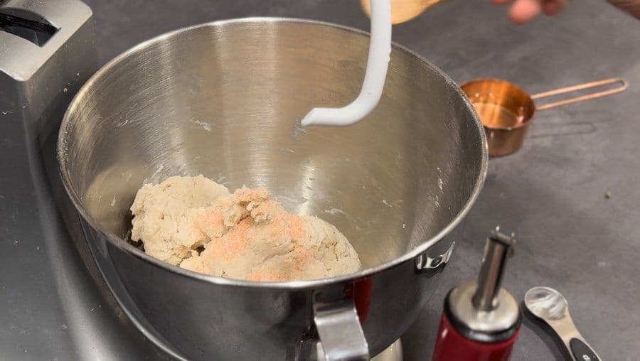 Pizza dough being mixed in a stand mixer bowl, showing a soft, pale dough with a flat beater attachment.