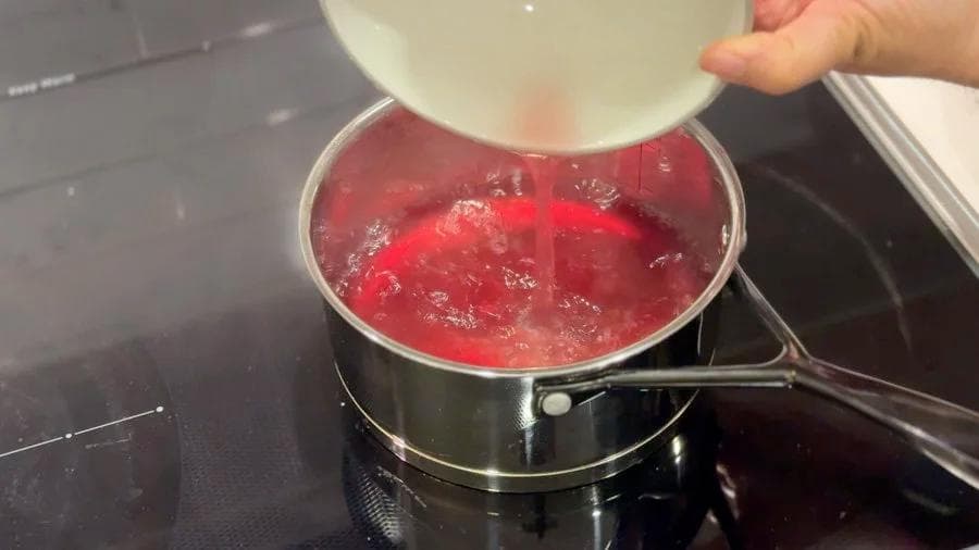 Pouring strained rose petal water into a pot of simmering red liquid for Gulabi Doodh.