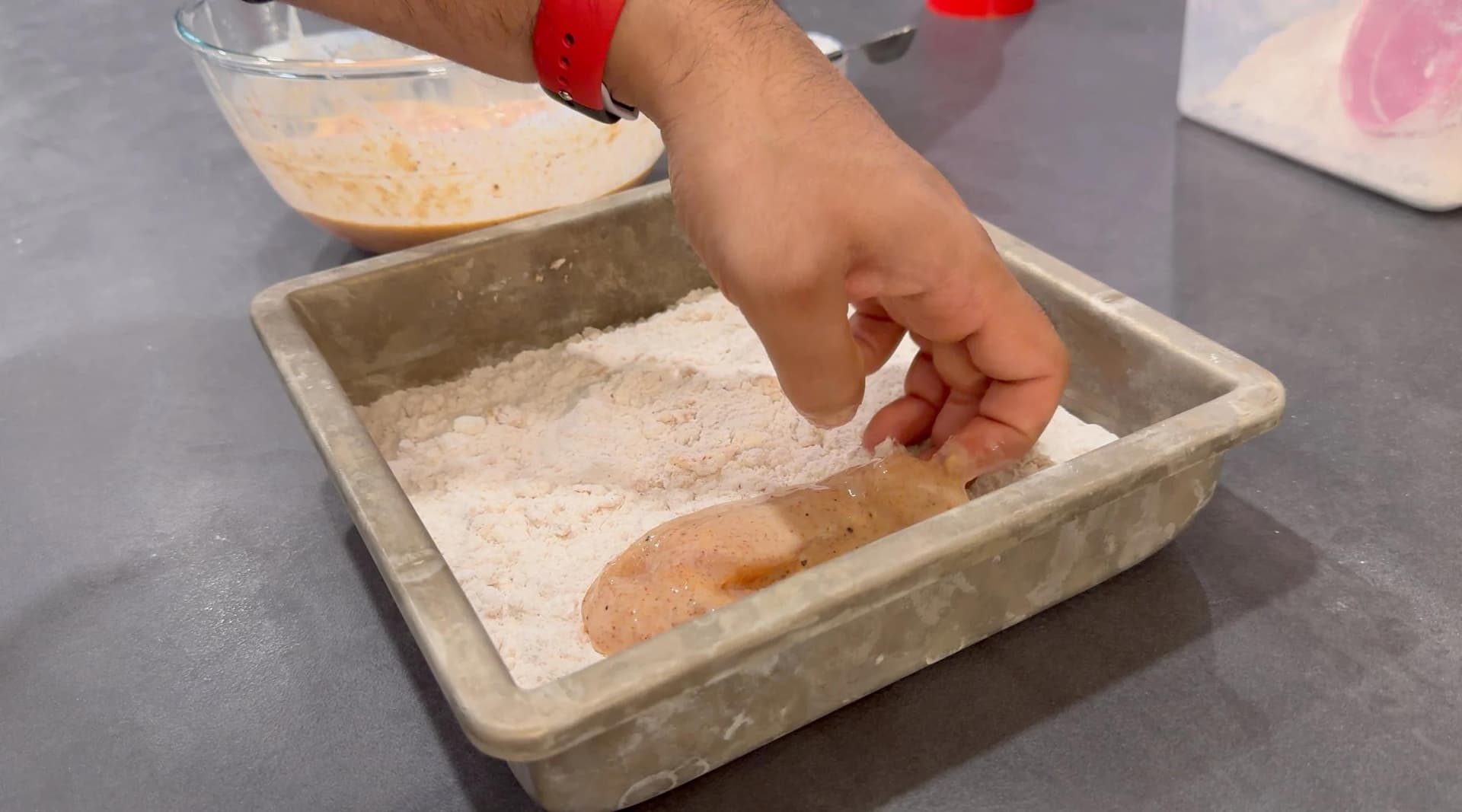 Hand coating chicken tender in seasoned flour mixture in a baking pan for fried chicken.