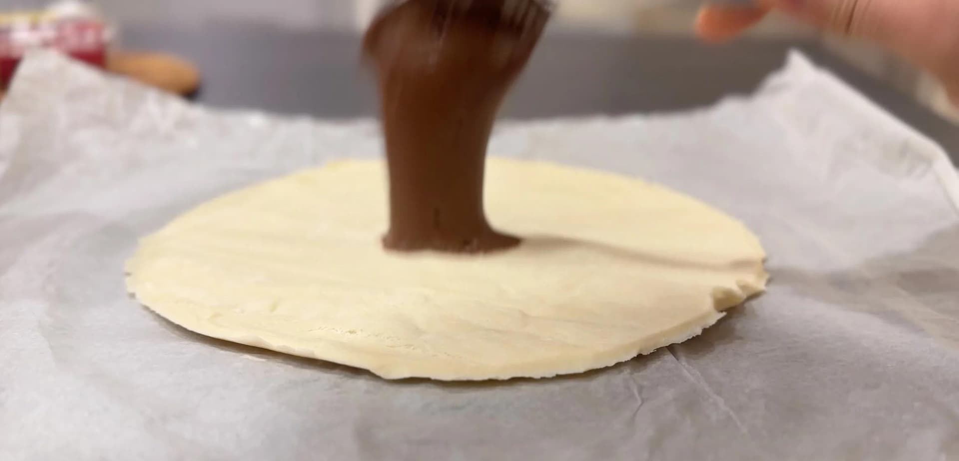 Hazelnut spread being spread on a circular puff pastry with a spoon, leaving a border around the edges.