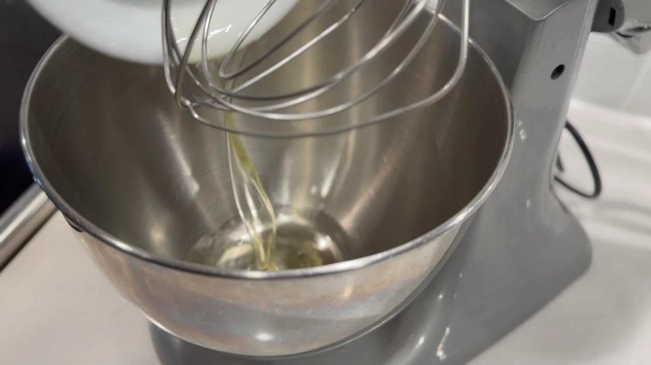 Whipped egg whites being lifted with a whisk attachment in a stand mixer, showing soft peaks forming in the stainless steel bowl.