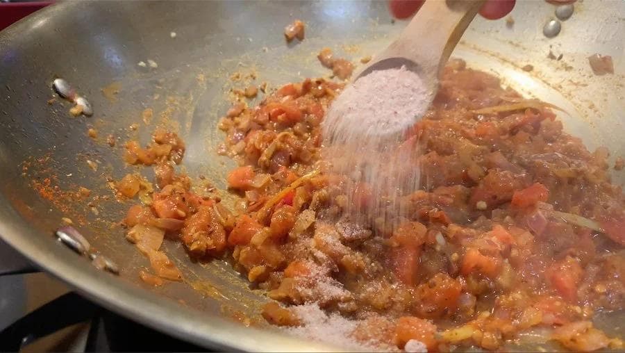 Sautéing diced onions and tomatoes in a wok with spices while stirring with a wooden spatula.