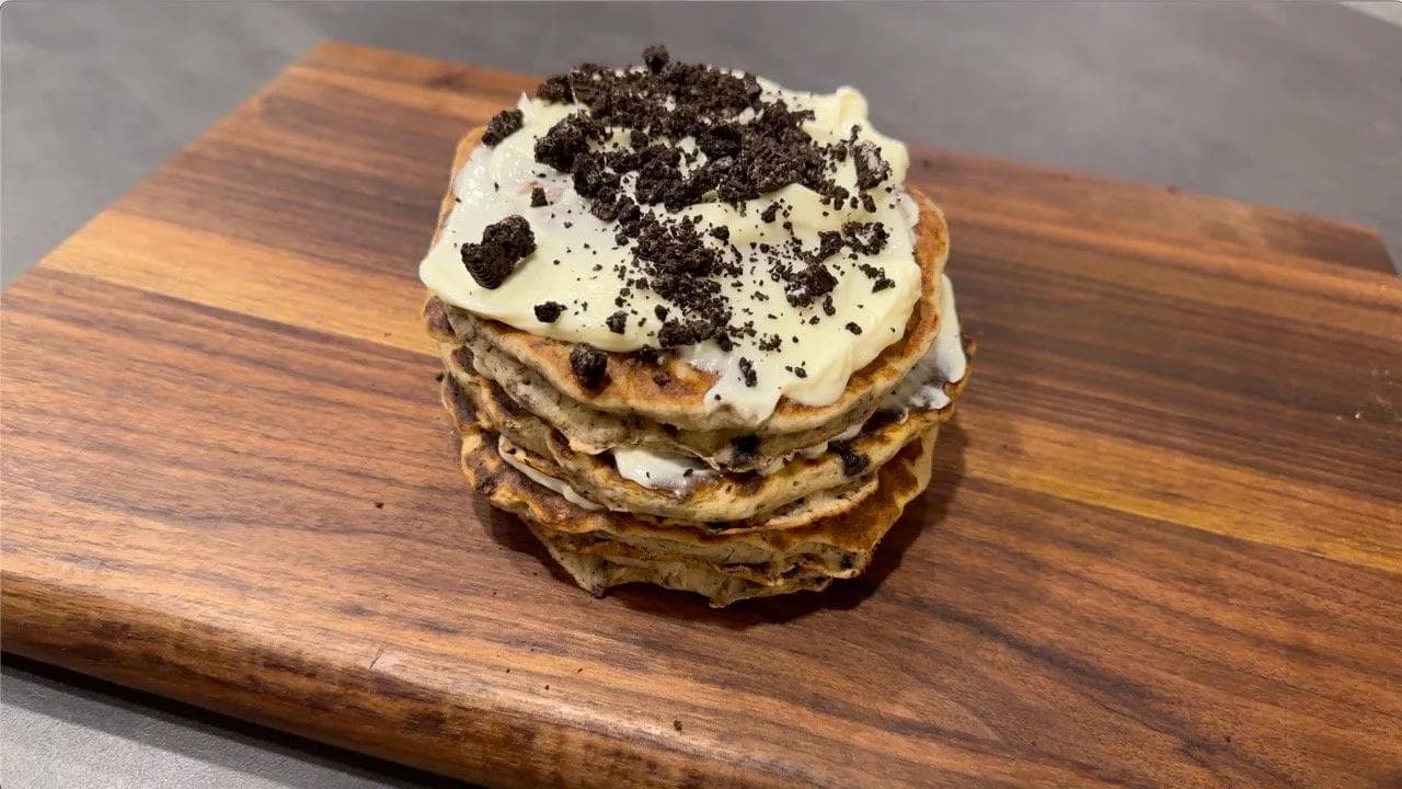 Stacked gluten-free Oreo cheesecake pancakes topped with whipped cream and crushed cookies on a wooden board.