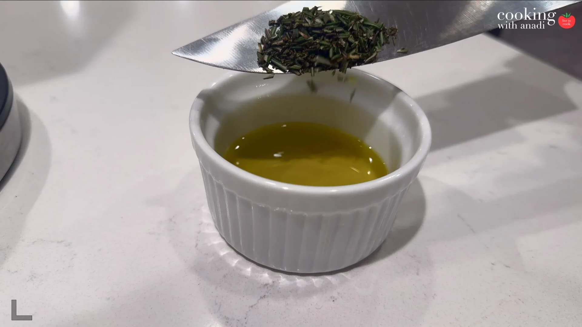 Dried rosemary being scraped off a knife blade into a small white ramekin of golden olive oil.