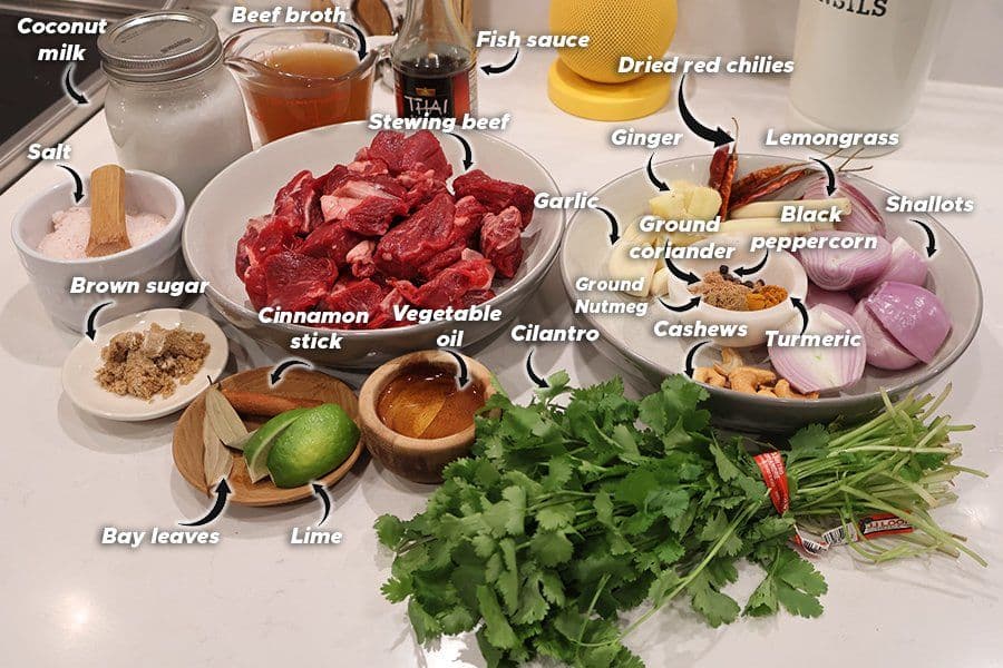 Ingredients for Balinese beef rendang laid out on a white surface including stewing beef, spices, herbs, coconut milk, and broth.