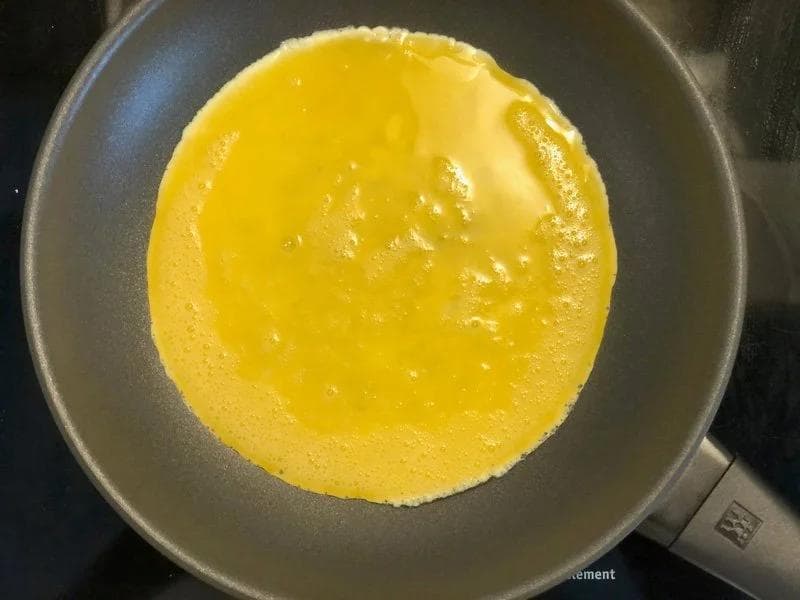 Whisked eggs cooking in a nonstick pan, forming a golden-yellow omelet base for an Indian spicy egg roll.