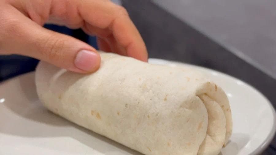 Hand holding a reheated breakfast burrito wrapped in parchment paper on a white plate.