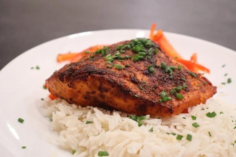 Blackened salmon fillet on white rice with green onions and orange carrot garnish on a white plate.