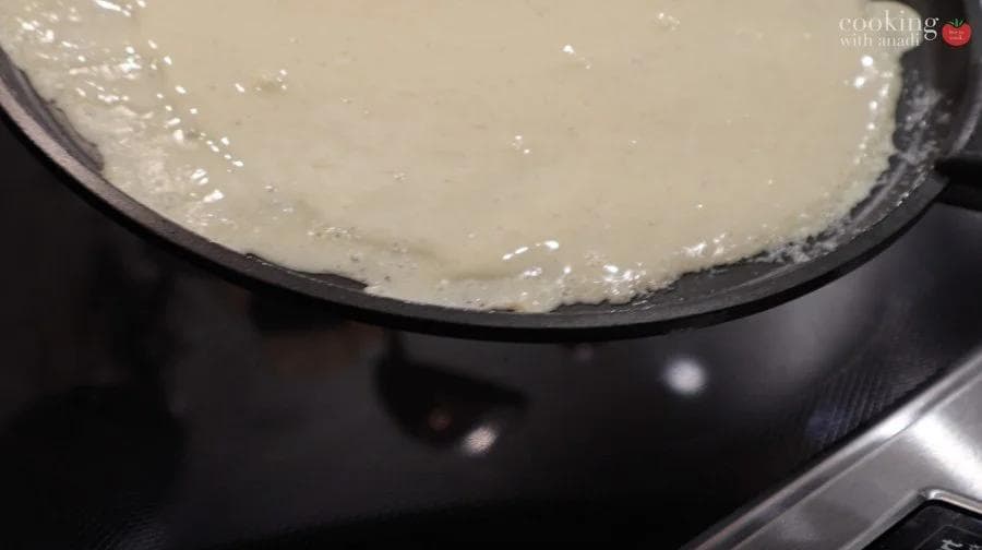 Crepe batter being cooked in a non-stick pan, showing a thin, light golden crepe.