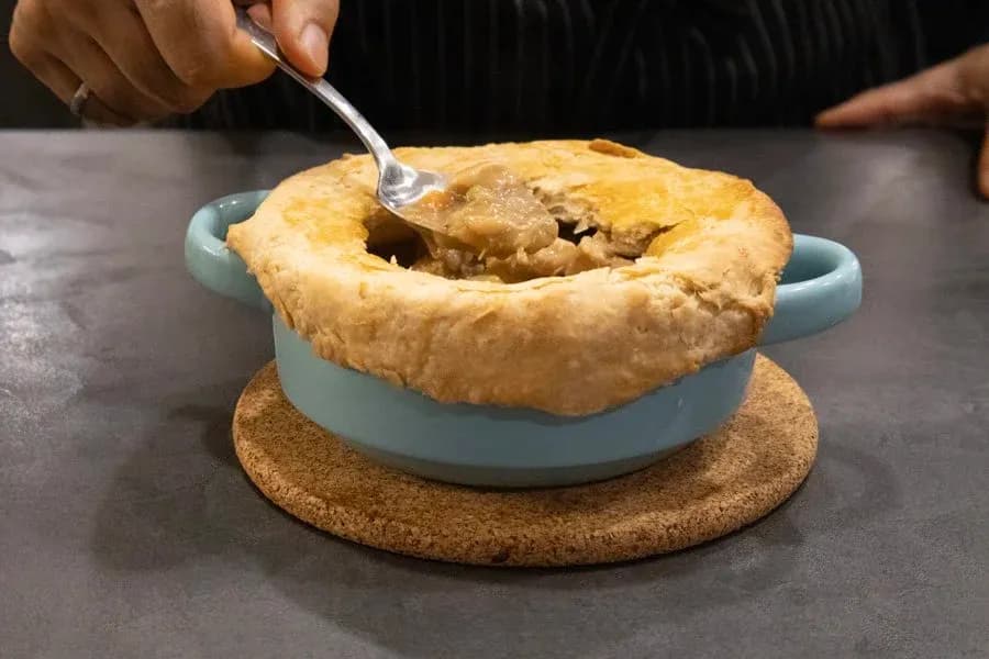 Spoon breaking into golden-brown Guinness chicken pot pie in light blue ceramic dish with cork trivet.