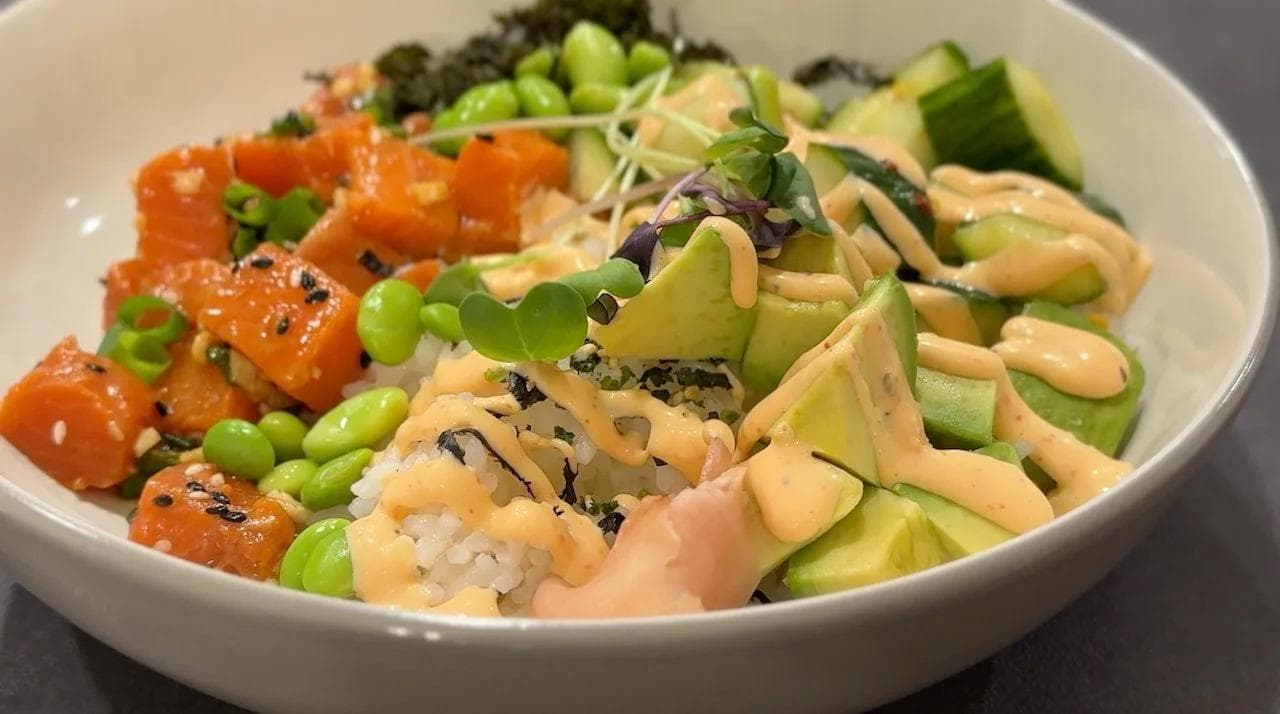 Assembled salmon poke bowl with rice, salmon cubes, avocado, edamame, cucumber, and furikake in a white bowl.