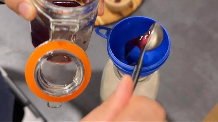Adding red food coloring to milk mixture in blue container while holding glass jar with orange ring.