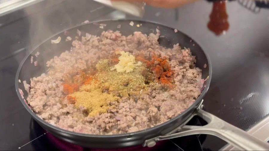 Ground beef cooking in a skillet with paprika, cumin, coriander, and minced garlic sprinkled on top.