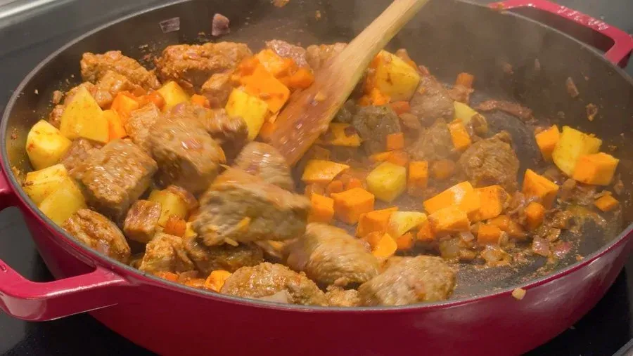 Portuguese beef stew with browned meat chunks and diced potatoes and carrots cooking in a red pot.