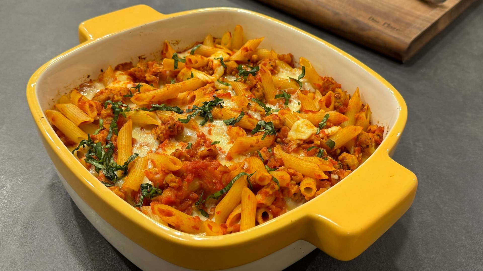 Cheesy baked penne with spicy Italian sausage in a yellow casserole dish, topped with melted cheese and fresh basil.