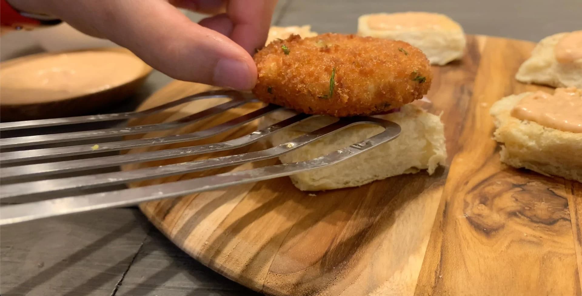 Golden-brown McAloo Tikki potato patty being placed on a fork over toasted bun with assembled burger ingredients on wooden board.