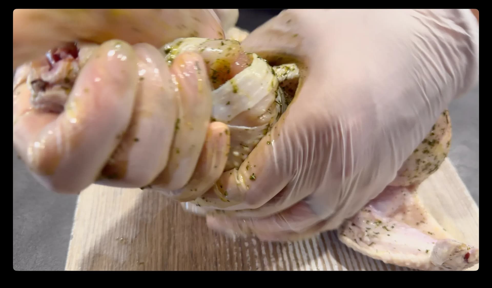 Gloved hands rubbing herb and oil paste over raw chicken on a wooden cutting board.