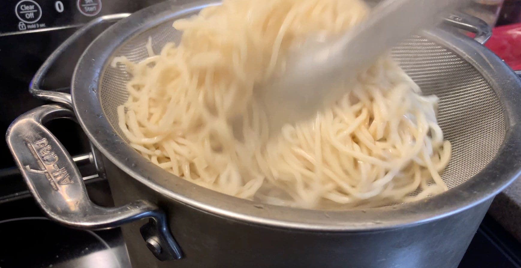 Cooked noodles being strained in a colander over a pot of boiling water.