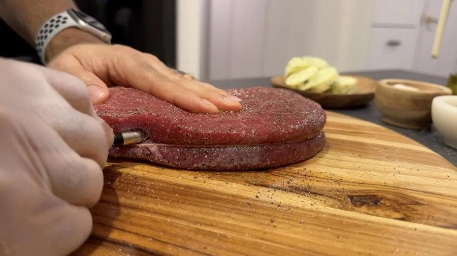Hand pressing meat thermometer into center of raw sirloin steak on wooden cutting board.