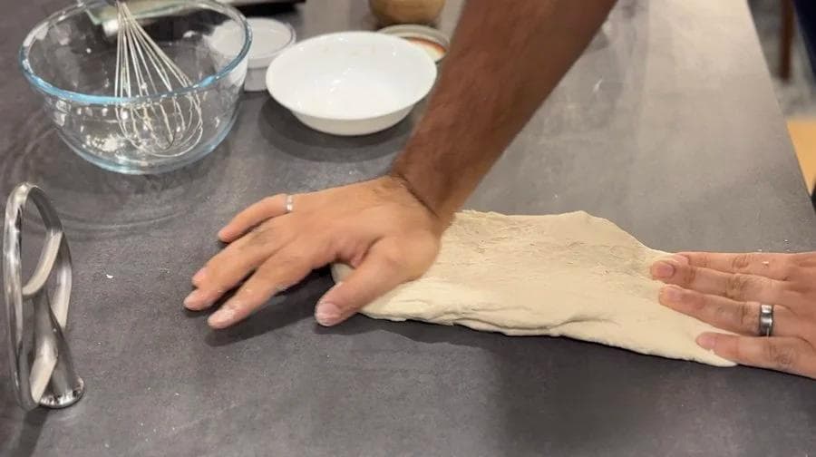 Hands stretching and folding dough on a dark work surface with a whisk and bowl nearby.