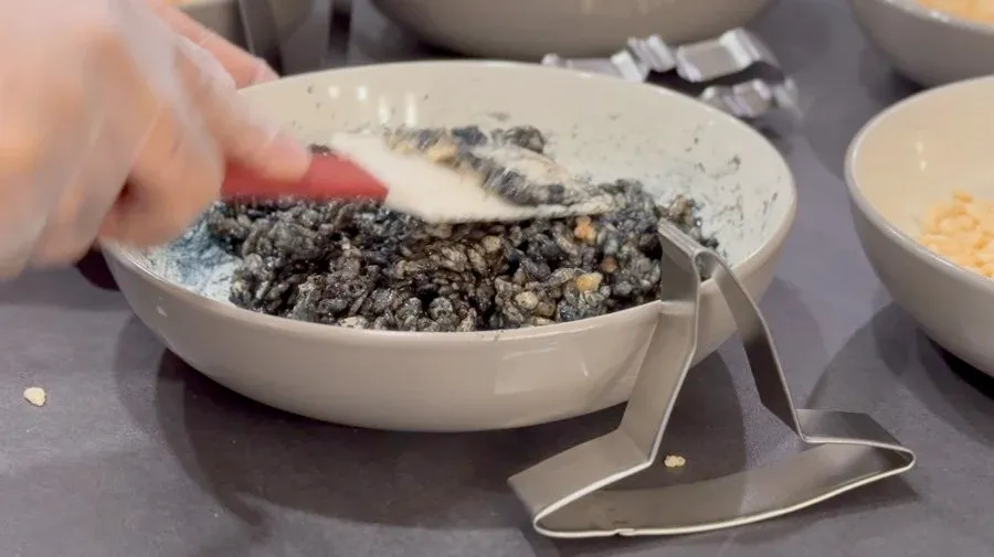 Hand spreading black Rice Krispies treat mixture in white bowl with bat-shaped cookie cutter.