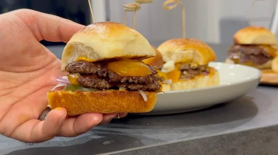 Hand holding a cooked burger slider on a dinner roll with melted cheese and red onion, with more sliders on a plate behind it.