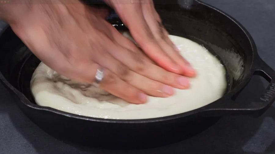 Hands pressing and dimpling pizza dough in an oiled cast iron skillet.