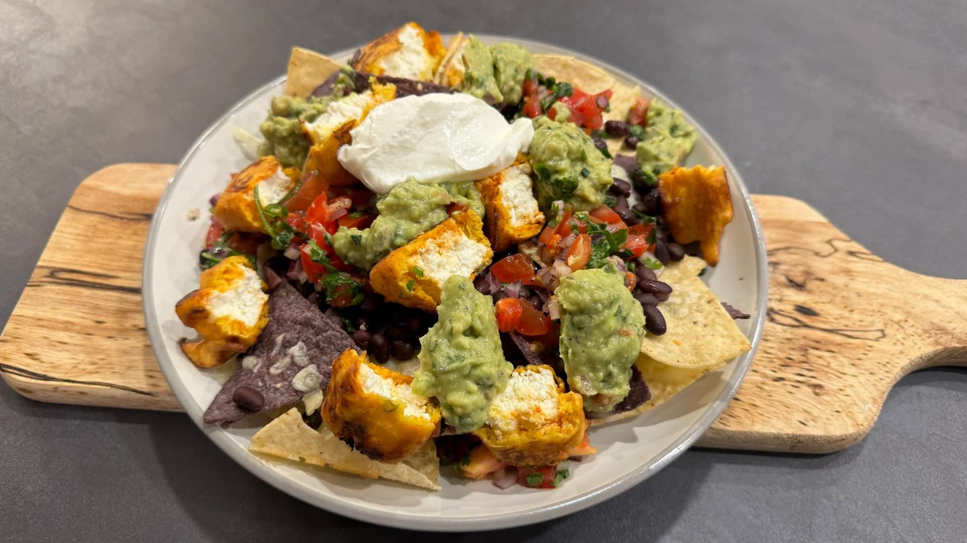 Paneer pakora nachos topped with guacamole, sour cream, diced tomatoes, and black beans on a white plate with tortilla chips.