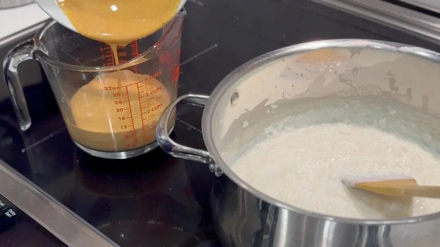 Creamy pumpkin rice pudding being stirred in a pot with a wooden spoon, with tempering mixture in a measuring cup nearby.