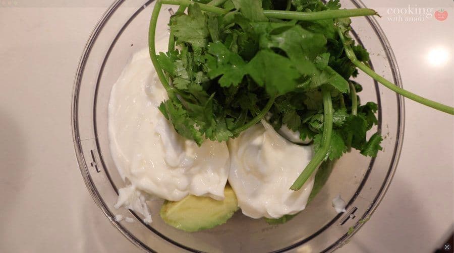 Food processor containing avocado, sour cream, and fresh cilantro ready to blend into crema sauce.