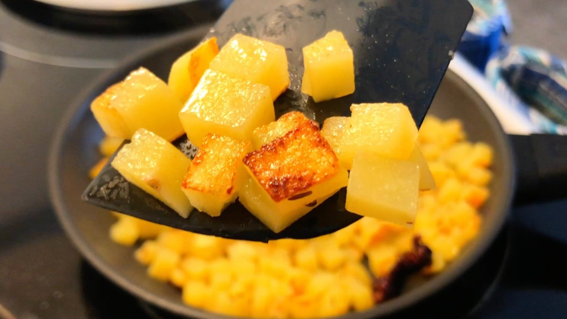 Cubed potatoes being turned with a spatula in a skillet, some pieces golden-brown, with corn kernels around the edges.