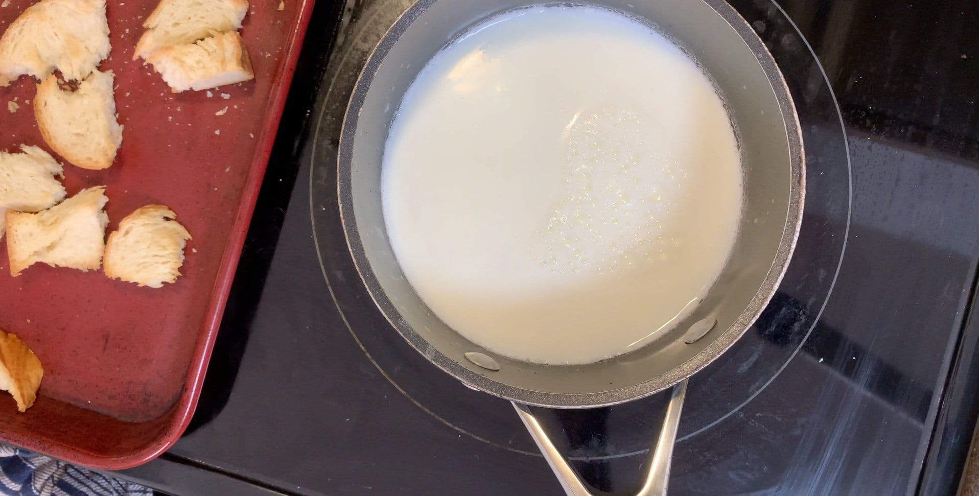 Simmering milk in a saucepan next to toasted brioche bread cubes in a red baking dish.