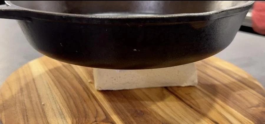 Cast iron pan pressing tofu block on wooden surface with paper towel underneath.