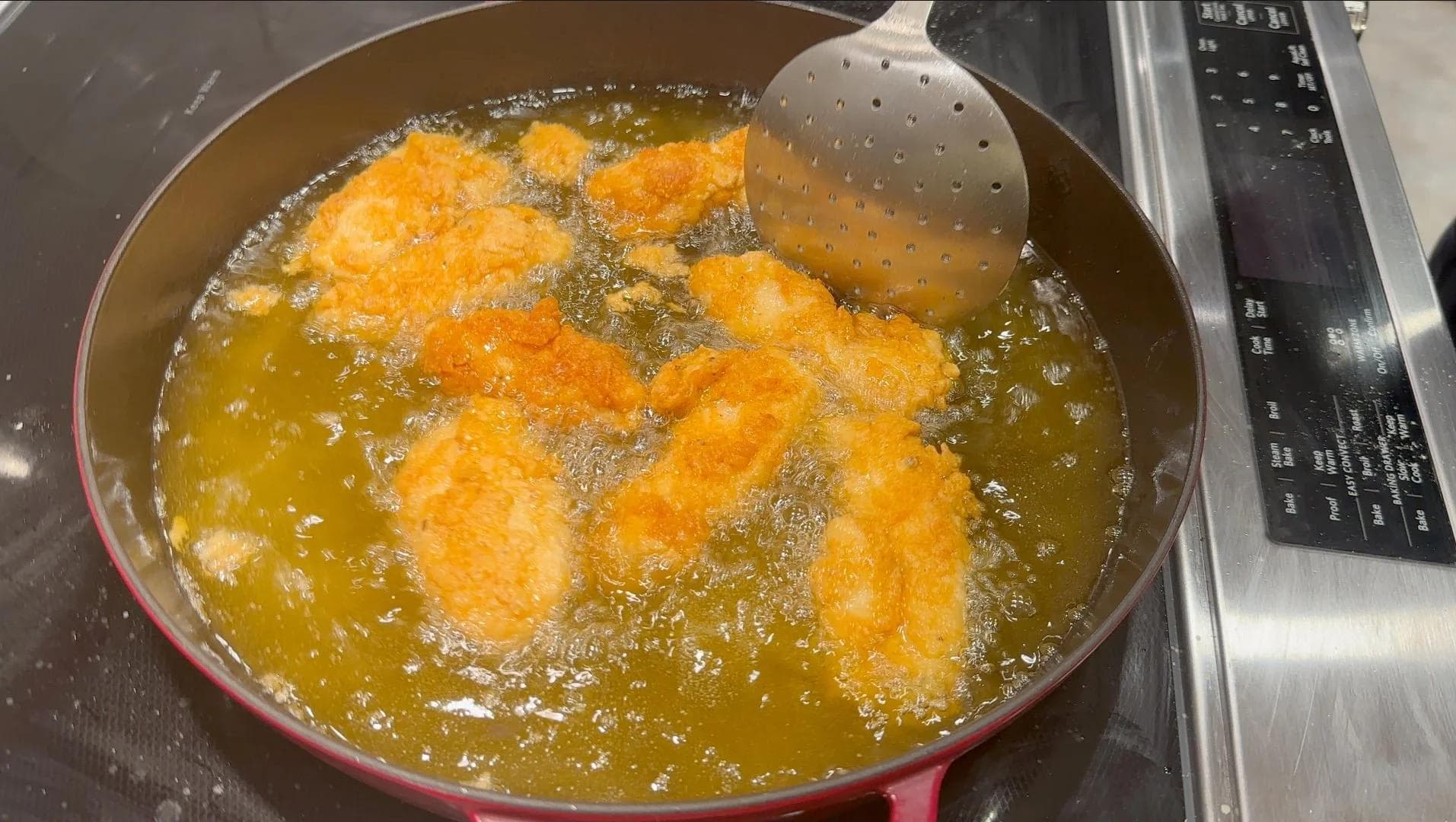 Golden-brown fried chicken tenders frying in hot oil in a red pan, being turned with a slotted spoon.