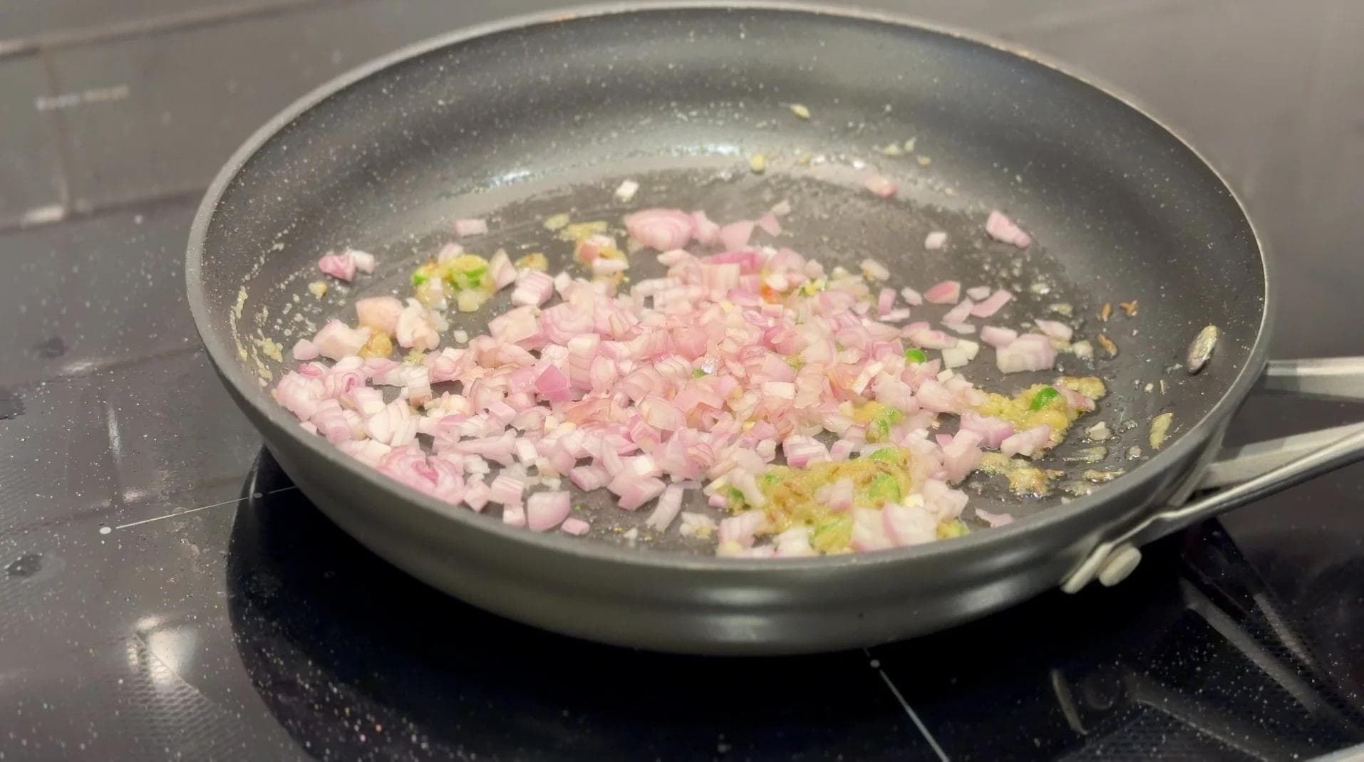 Diced shallots and green chillies sautéing in oil in a dark pan on a stovetop.