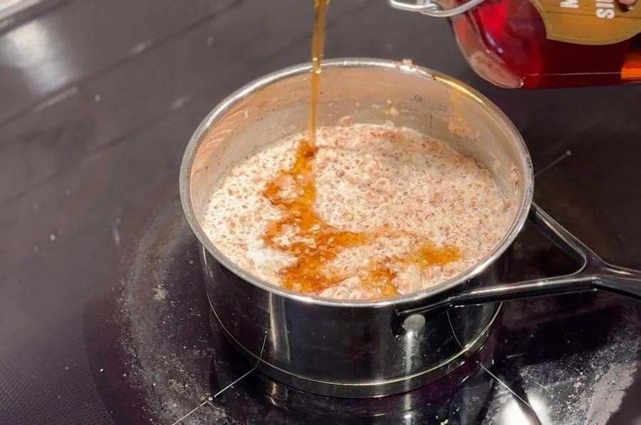 Maple syrup being poured into a pot of cooked cracked wheat porridge while stirring on the stovetop.