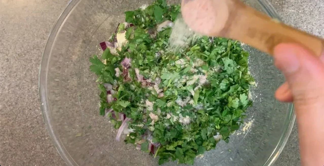 Mixed dry ingredients for besan chilla including chickpea flour, chopped herbs, onions, and spices in a glass bowl.