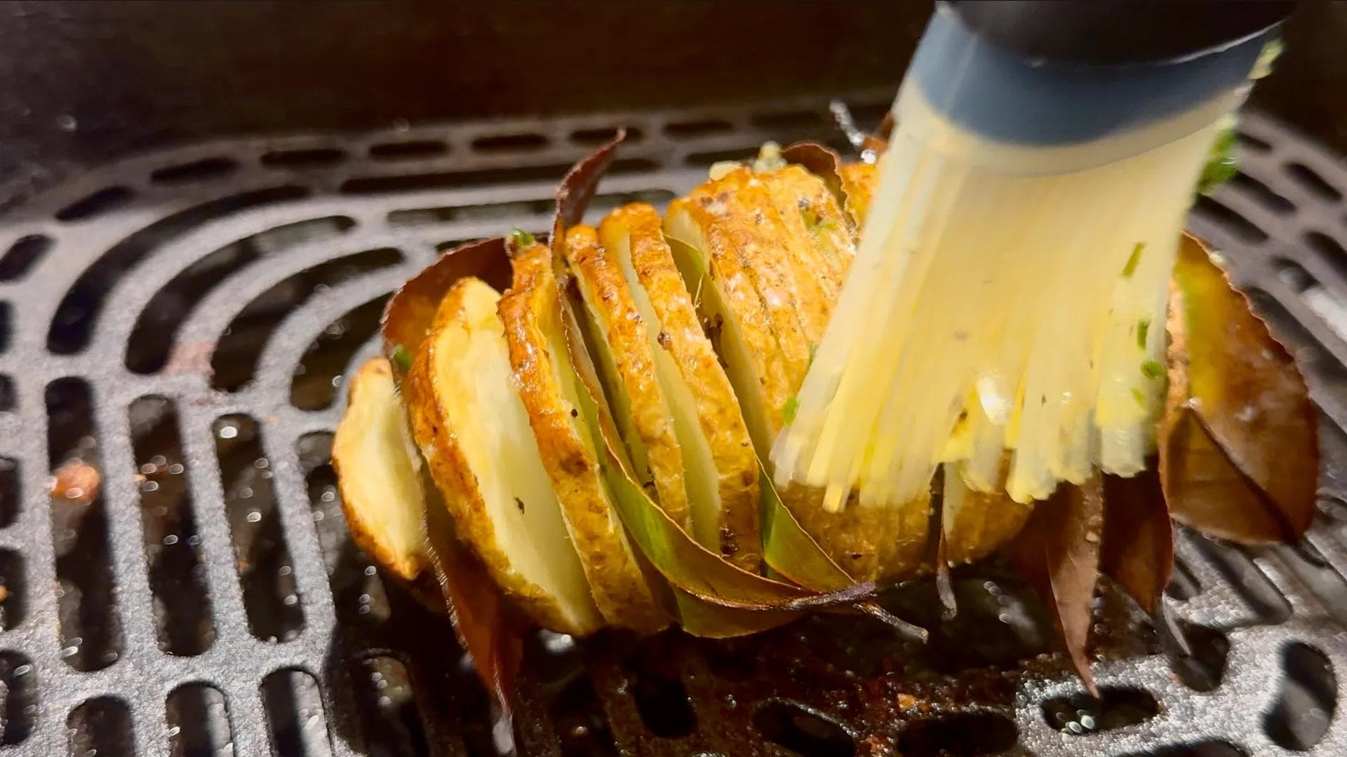 Hasselback potato being brushed with melted butter in air fryer basket during cooking.
