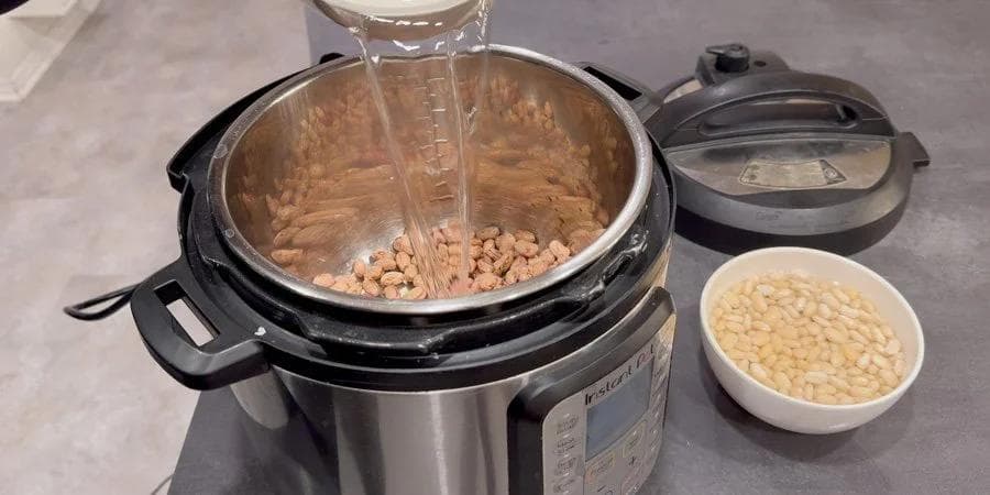 Water being poured into Instant Pot with soaked pinto beans and a bowl of Great Northern beans nearby.