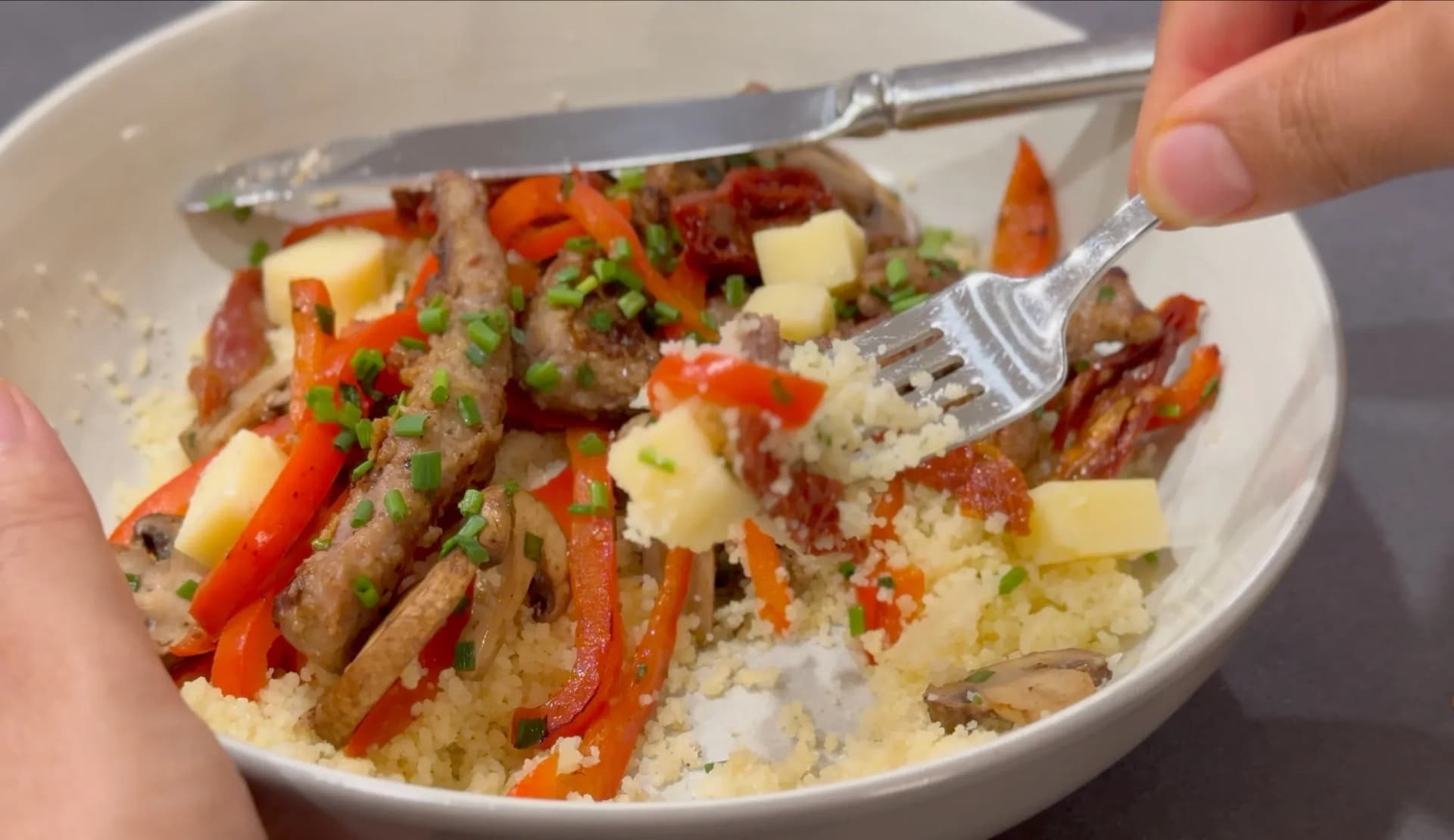 A fork lifts a bite of beef and vegetable couscous topped with cheddar cheese cubes and fresh chives from a white bowl.