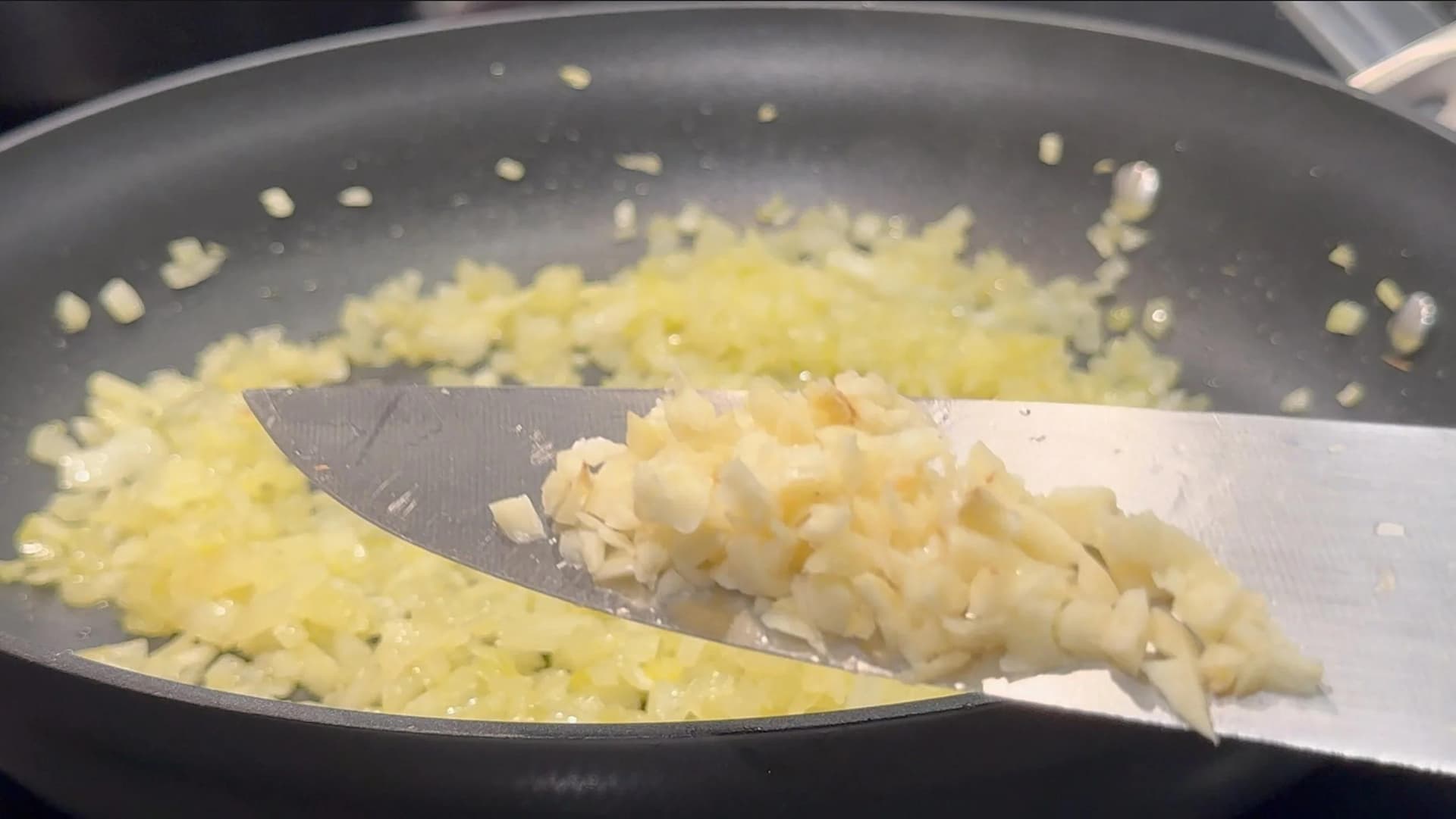 Diced onions and garlic sautéing in a dark non-stick pan, being stirred with a metal spatula.