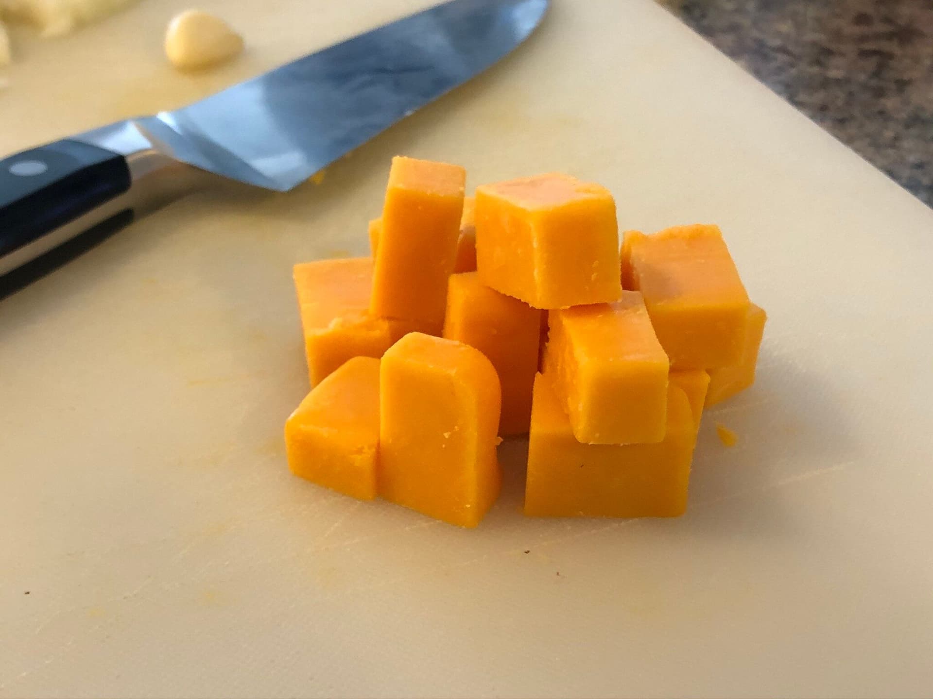 Cubed cheddar cheese on a cutting board next to a chef's knife during mac and cheese preparation.