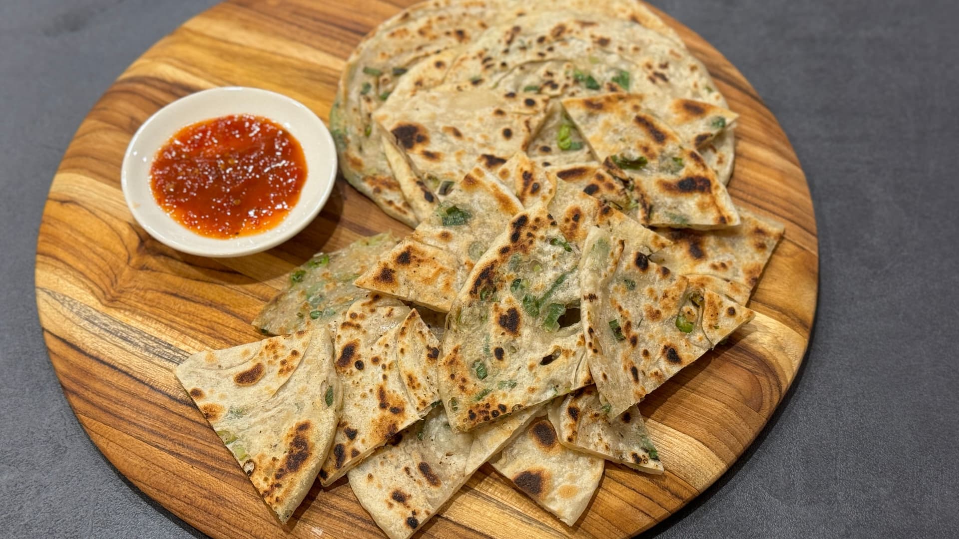 Crispy golden Chinese scallion pancakes cut into triangles on a wooden board with a small bowl of red chili sauce.